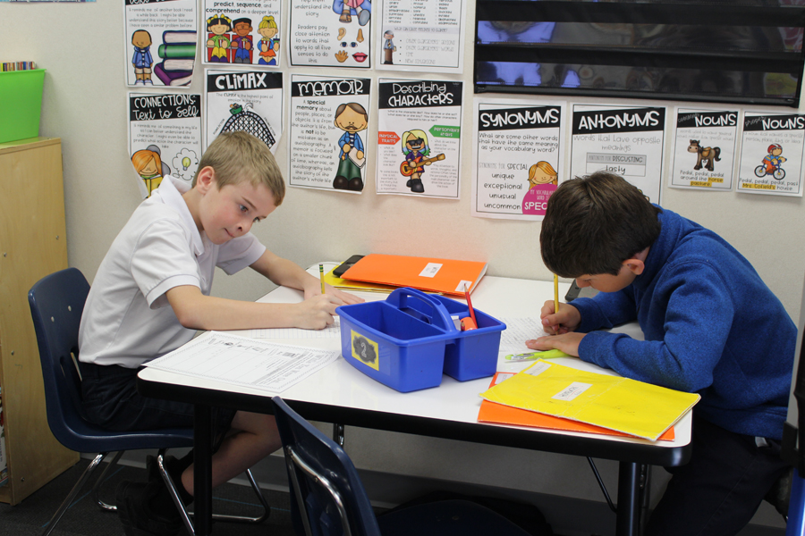 Two third-grade boys sit at table in classroom, writing on paper