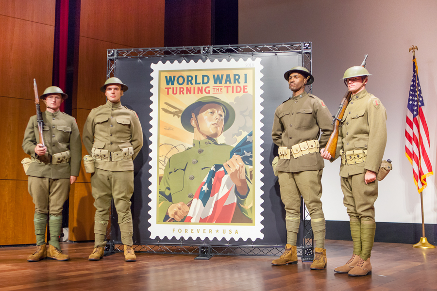 Four men in World War I era uniforms stand near stamp poster