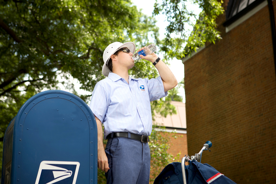 Letter carrier stands next to blue collection box, drinking bottled water