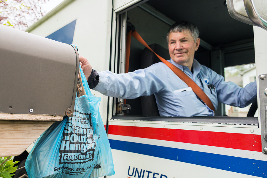 Letter carrier sits in delivery vehicle and picks up bag marked Stamp Out Hunger at mailbox