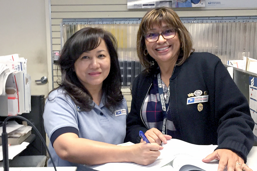 Two smiling women stand at Post Office counter filling out form