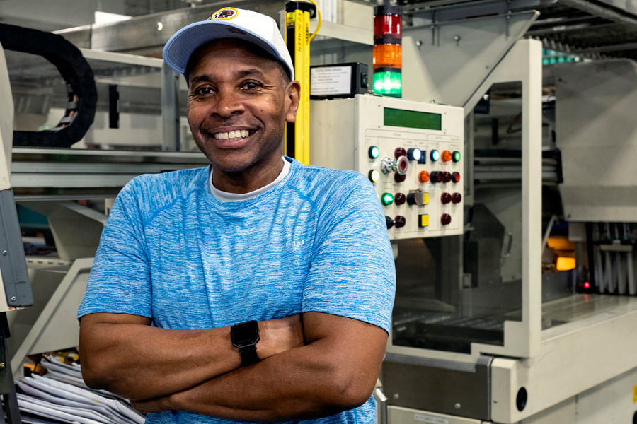 Smiling worker stands near machinery on postal plant floor