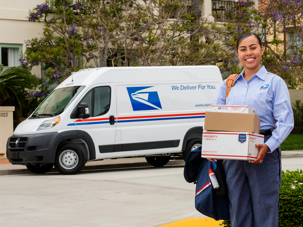 Smiling letter carrier holds packages near postal delivery vehicle