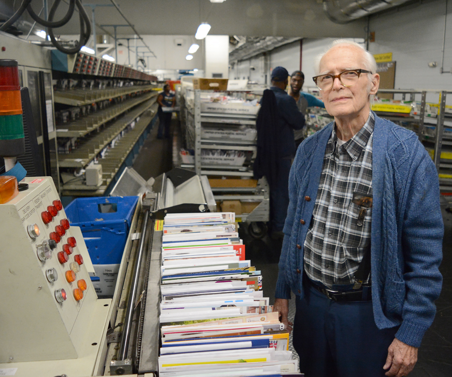 Man stands on floor of processing plant, near mail sorting equipment