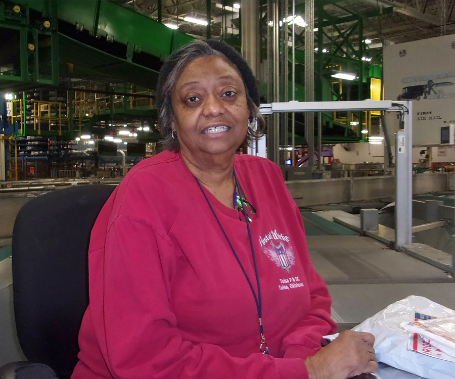 Smiling postal worker sits near processing machine on workroom floor